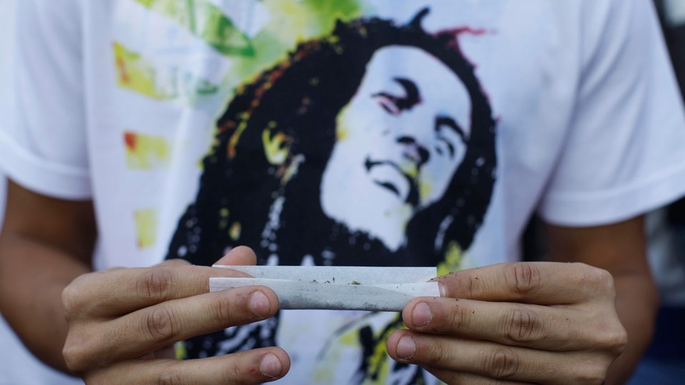 Ein junger mann dreht sich einen Joint. Im Hintergrund ist sein T-Shirt imit Bob Marley Aufdruck zu sehen.