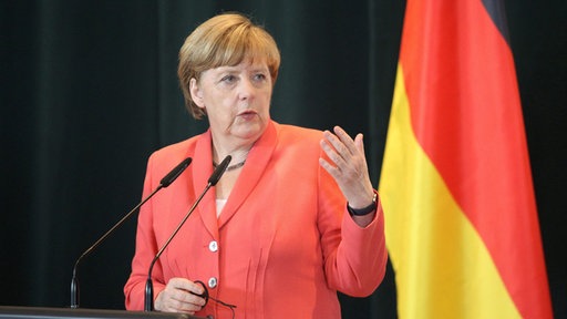 Angela Merkel u crvenom sakou, ispred mikrofona, za govornicom