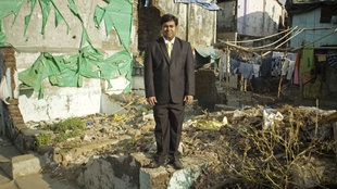 Filmszene "India" aus der Doku "I want to see the manager", ein Man im Anzug steht vor kaputter Hausfassade
